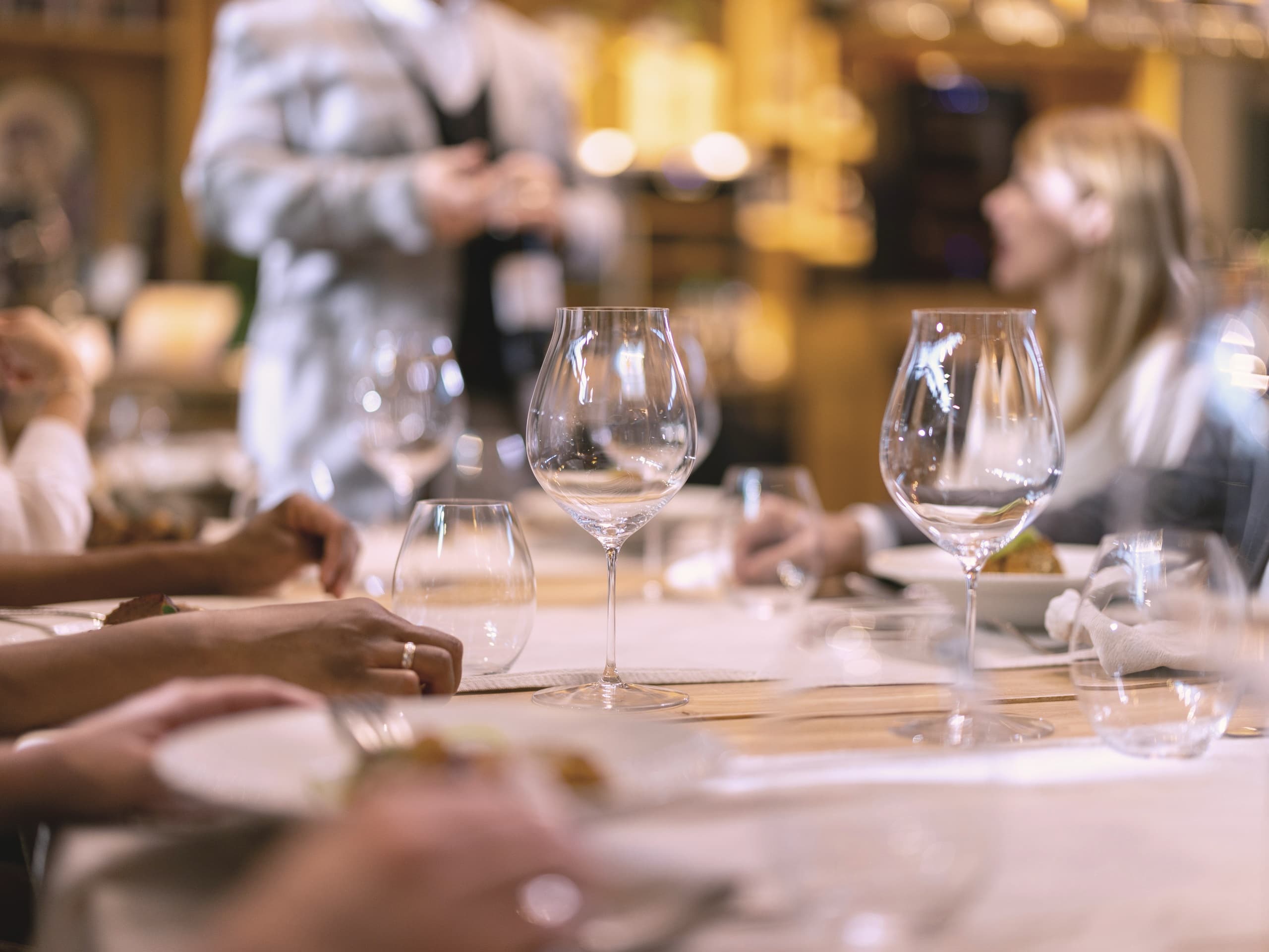 Gäste an einer langen Tafel, Gläser und Hände im Vordergrund